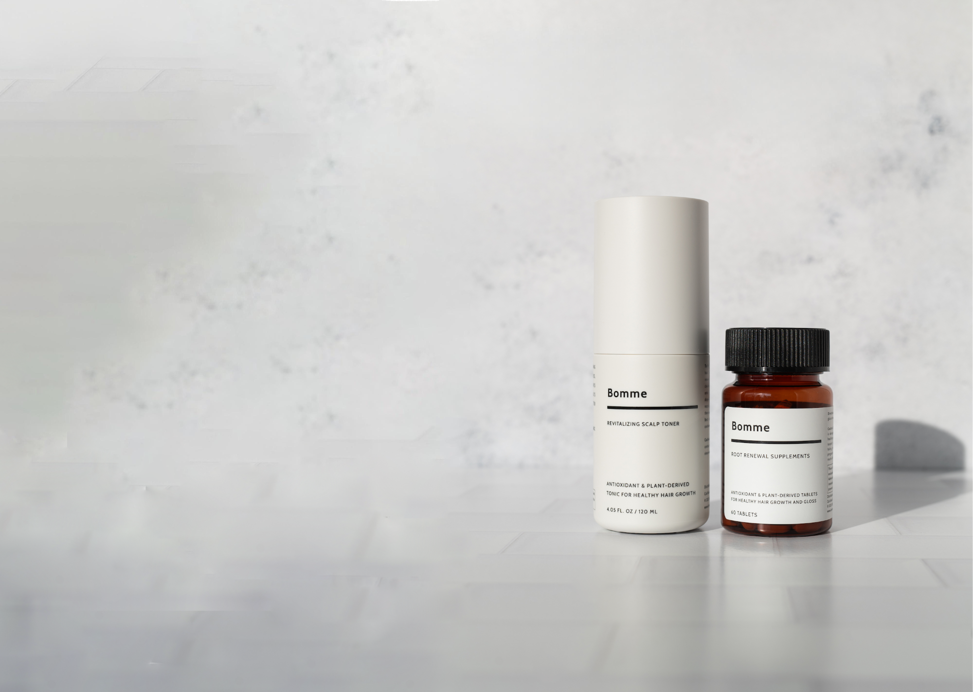 Product display of Bomme Root Renewal Supplements and Revitalizing Scalp Toner on a tiled surface with a light textured background, positioned for hero image layout with open space for text.