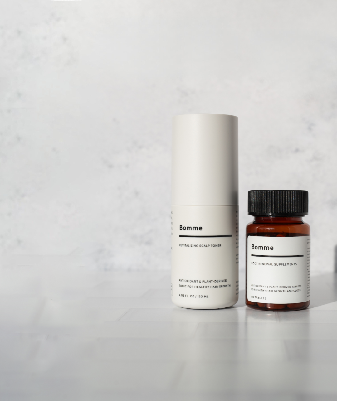 Product display of Bomme Root Renewal Supplements and Revitalizing Scalp Toner on a tiled surface with a light textured background, positioned for hero image layout with open space for text.